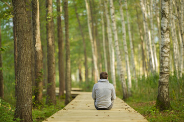 Young adult man sitting on wooden trail at birch tree forest. Looking far away. Spending time alone...