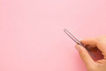 Young adult woman fingers holding tweezers on light pink table background. Pastel color. Closeup....
