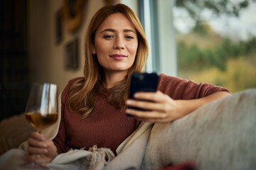 A smiling blonde girl enjoying a glass of wine while using a mobile phone.