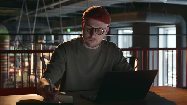 A young man of 30 years old is a freelancer working on a laptop in a dark room of a loft cafe. A freelancer makes notes in a notebook with a pen while working on a laptop. High quality 4k footage
