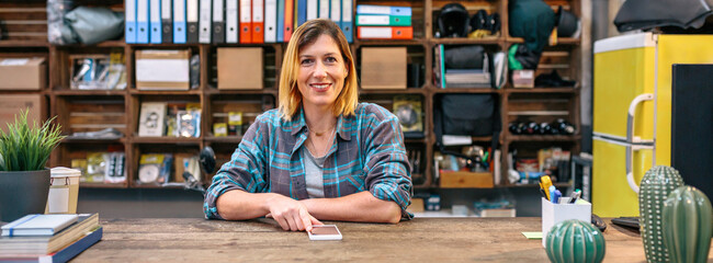 Portrait of smiling blonde young female employee looking at camera behind of industrial shop counter. Banner of happy woman with freckles and plaid shirt working in store selling motorbike spare parts - obrazy, fototapety, plakaty