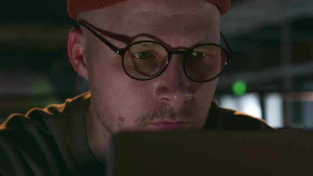 A close-up portrait of a young freelancer in round glasses and a hat in a dark loft cafe. A man is concentrating on a computer in a dark room. High quality 4k footage