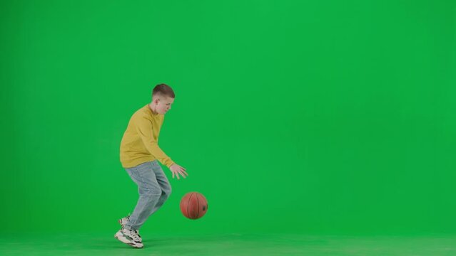 Portrait of boy on chroma key green screen. School boy kid in casual clothing walks through the frame and plays with basketball ball.