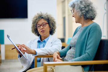 Female doctor going through medical paperwork with her senior patient in waiting room.