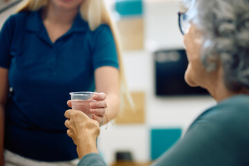 Close up of nurse giving glass of water to senior patient in waiting room at doctor's office.