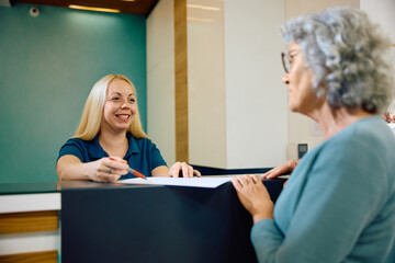 Happy nurse and senior woman talk while filling medical data at medical clinic.