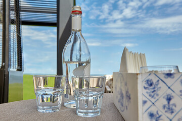 A glass bottle of drinking water and two glasses on the table of an outdoor summer cafe against a...