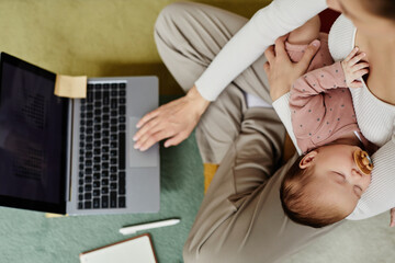 Top down view at adorable baby girl with pacifier in mouth sleeping peacefully in arms of unrecognizable mom using computer on floor at home, copy space
