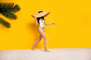 Full length photo of cheerful positive lady wear swimsuit sun cap walking beach empty space isolated yellow color background
