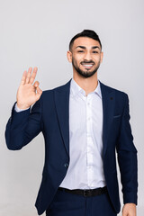 Handsome young turkish man in smart clothes showing okey looking at camera smiling standing over grey background in studio isolated.