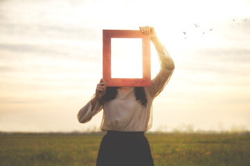 woman holds an empty frame that makes her face disappear reflecting the sky, abstract concept