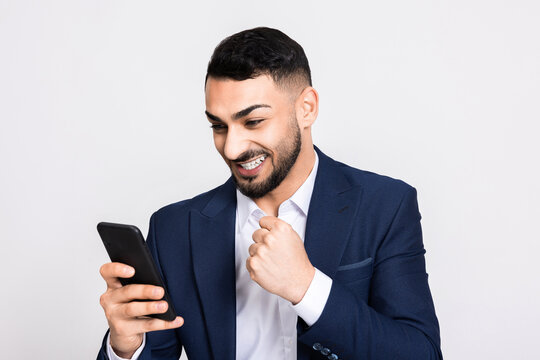 Cheerful attractive turkish man in smart suit holding new modern smartphone observing photos i phone chatting messaging with friends family girlfriend gambling trading online celebrating victory.