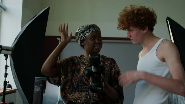 Young Caucasian male model discussing photos on digital camera with Black female photographer after fashion shoot in professionally equipped studio