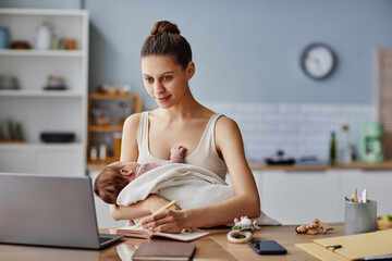 Smiling young Caucasian mom looking at laptop and writing in notebook at table while holding sleeping newborn, copy space - Powered by Adobe