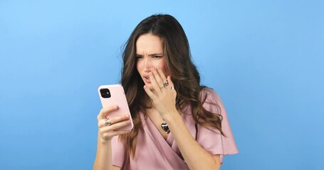 Close up young girl sad while read a message about broke up. Sad young woman isolated on blue background looking at smartphone screen and be melancholy after receiving bad news. Bad mood.