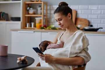 Side view at happy Caucasian mother sitting in kitchen using smartphone while holding napping baby in blanket, copy space - obrazy, fototapety, plakaty