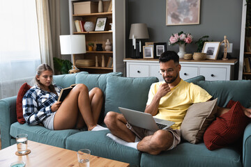 Happy young man working on sofa with laptop near woman with book lying on sofa in living room at...
