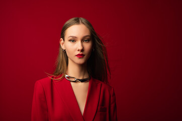  Fashionable confident beautiful woman with red lips, eyeliner makeup, wearing suit blazer, chunky metallic necklace, posing on red background. Close up studio portrait. Copy, empty space for text