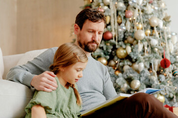 father and little daughter sit on floor near christmas tree at home, and read fairy tale book...