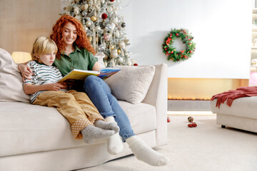 mother and little son sit on couch near christmas tree at home, and read fairy tale book together, smile and laugh at funny stories