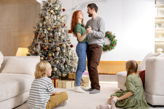 Mom And Dad Dancing In Living Room Near Decorated Christmas Tree, Kids Sit On Floor And Clapping Their Hands. Family Time Together, Celebrating, Smiling And Laughing