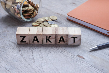 Wooden cubes with text on ZAKAT on the wooden table. Coins flowing out from the glass container, and notebook at the back. Business and zakat concept