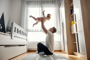 Father is sitting on the floor in nursery room and tosing his daughter in the air during their playtime. - obrazy, fototapety, plakaty