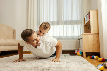 Father is doing pushups at home while the boy is on his backs and encourages him.