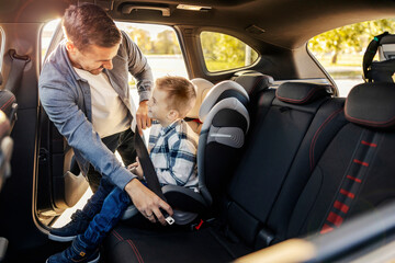 A happy dad is putting his son in his car seat. They are getting ready for travel.