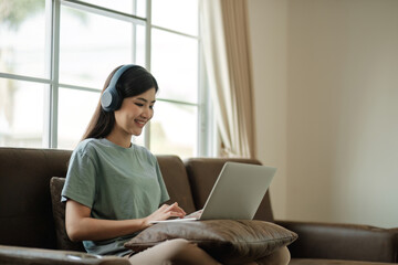 Young female student wearing headphones sits intently and happily studying online on her laptop on the sofa in the living room at home.