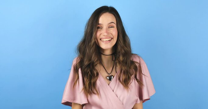 Happy friendly young woman waves palm in hello gesture welcomes someone with hospitable expression expresses positive emotions standing isolated bright blue background. Hi to all of you.