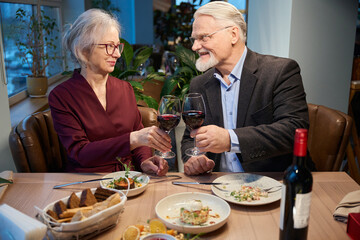 Aged woman and man enjoying festive dinner clinking wineglasses on Christmas