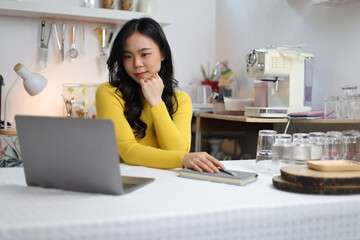 Young Asian woman working on laptop in kitchen at home, she is taking cooking class online and...