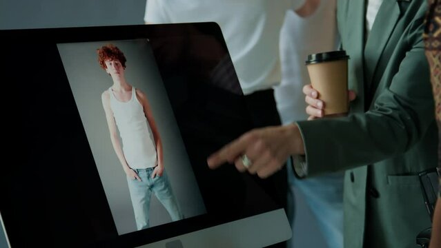 Female creative director holding coffee and discussion photo on computer with professional photographer during fashion shoot in studio, stylist working with model in background