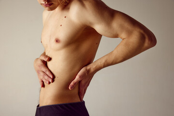 Cropped image of muscular shirtless male body against grey studio background. Young man holding hands on lower back. Pains, injury. Concept of men's beauty, health, body care, sportive lifestyle