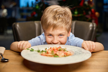Cheerful little boy looking at meal in restaurant while celebrating Christmas