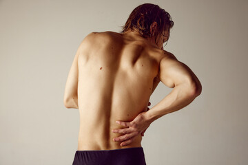 Lower back pains. Rear view of shirtless young man suffering from back pains against grey studio...