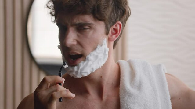 Close up funny happy Caucasian millennial man singing in shaving razor with foam on beard foamy bearded with shave gel cosmetic sing song having fun in morning bathroom listening music beauty routine