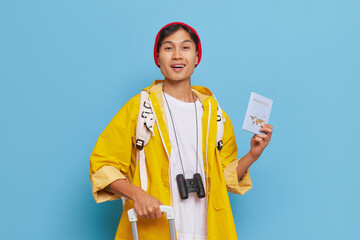 Happy tourist in yellow raincoat stands over blue background going to travel, journey time concept,...