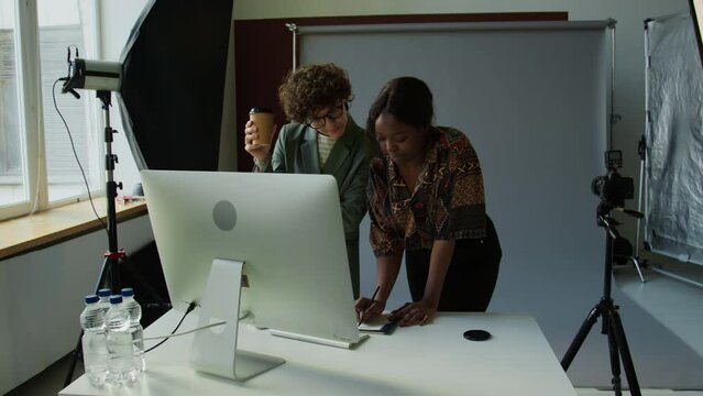 Female art director and photographer standing behind desk, looking at computer screen and discussing results of photo shoot in studio