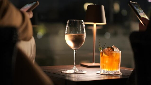 Primo piano su un calice di vino e su un Cocktail illuminati da una lampada da tavolo.
Due persone scattano foto con lo smartphone.
