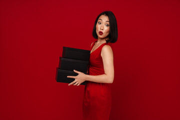 Surprised woman holding gift boxes and looking at camera isolated over red background