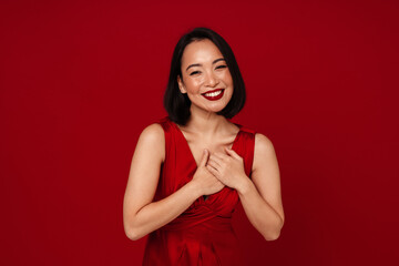 Lovely asian woman smiling and holding her hands on chest isolated over red background