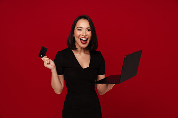 Naklejka premium Excited woman using laptop computer and credit card for online shopping isolated over red wall