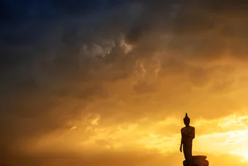 Fototapeten Stand big Buddha Statue in silhouette sun set Light background in park of Thailand temple.Yellow orange light silhouette dark shadow of image Buddha statue stand Buddha tall walk in sun light cloud. © BESTIMAGE