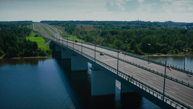 A modern asphalt highway winding away into the distance, winding through green meadows and forests, spanning over a bridge leading over a river. Drone shot