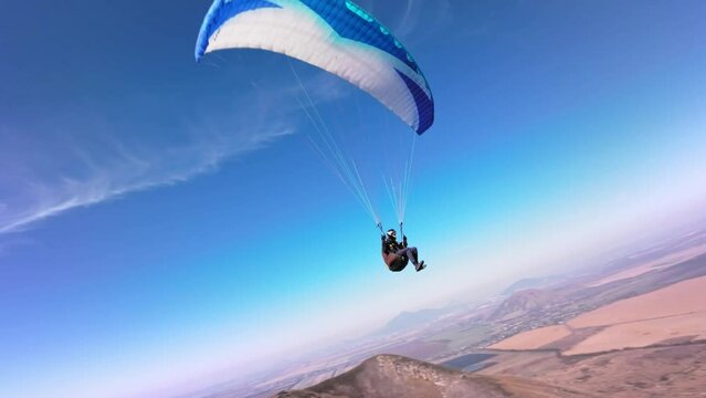 A paragliding athlete flies on a paraglider near the slope of a small mountain near a village on a sunny autumn day. Aerial view shooting with FPV drone quadcopter in acro mode