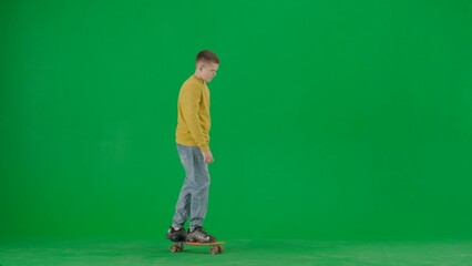 Portrait of kid boy on chroma key green screen. Schoolboy in jeans standing riding on skateboard through the scene, happy face expression. Full body shot