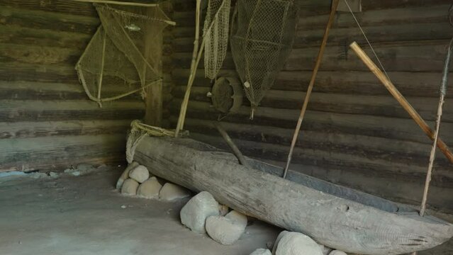 Old narrow wooden fishing boat stands on stones in storage facility, no people. Ancient nets and fishing gear hang on wooden barn wall in authentic village.