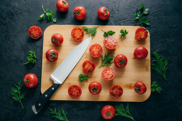 Top view of red sliced tomatoes on wooden chopping board. Sharp knife near. Green parsley and dill....